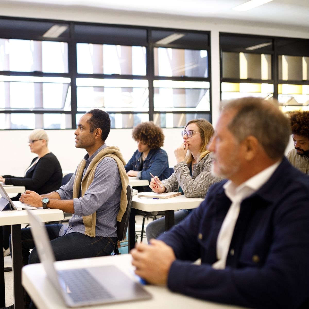 Diverse adults attentively sitting at desks in a modern classroom with laptops and notebooks.
