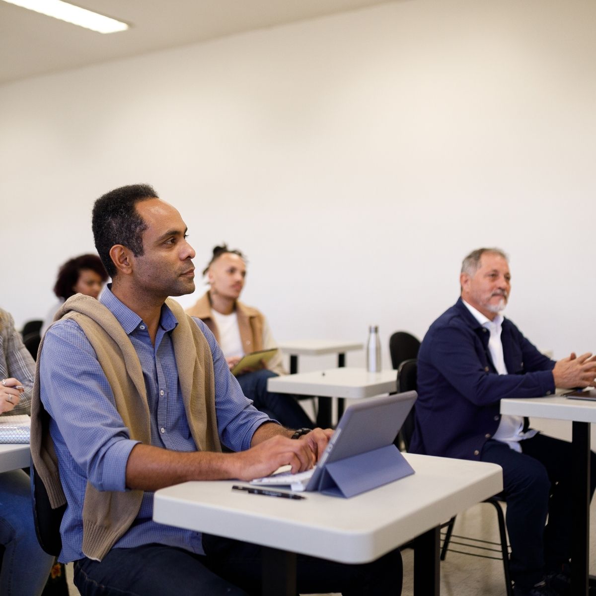 Diverse adults attentively sitting in a classroom with tablets and notebooks on desks.