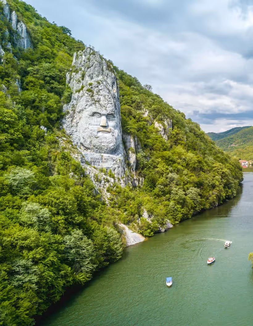 Stone sculpture of a large face carved into a green forested cliff beside a river with three small boats on the water.