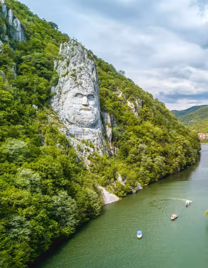 Rock formation with a large carved human face along a green riverbank with boats on a calm river.