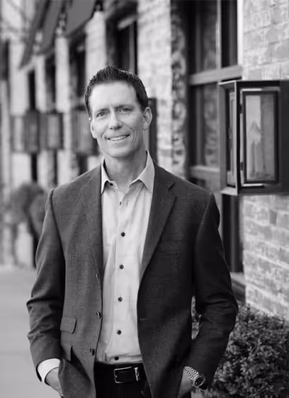 Smiling man in a suit jacket and dress shirt standing outdoors near a brick wall with lantern-style lights.