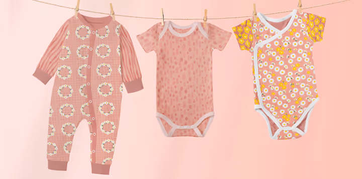 Three pint baby onesies on a pink background. The onesies feature the Summer Lawn pattern collection in the Sunrise colorway, with white English daisy and yellow buttercups on a peach pink background color.