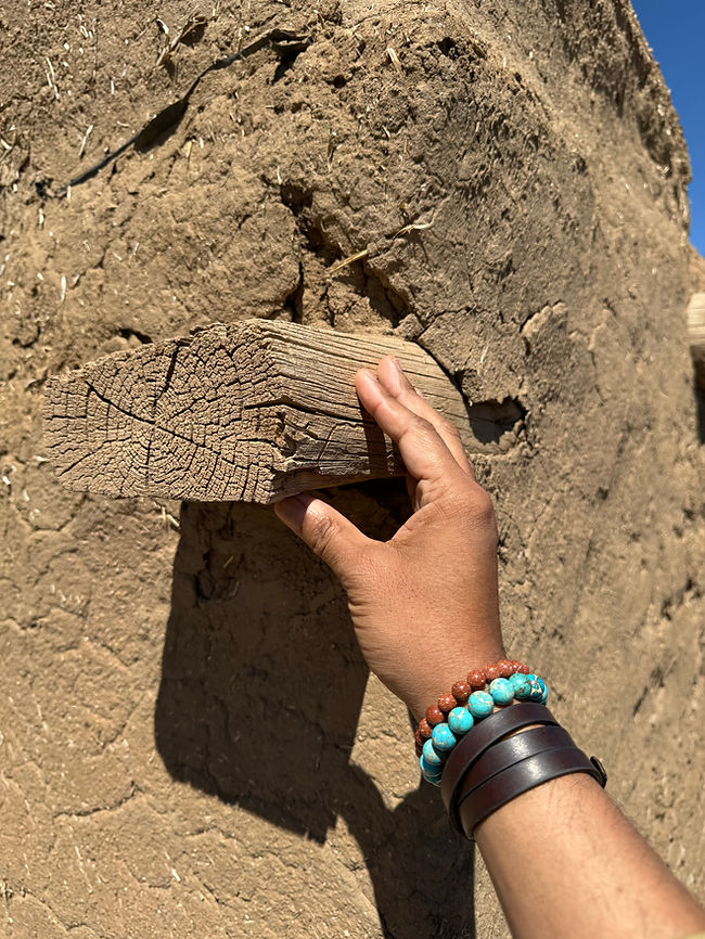 Hand touching weathered adobe wall and wooden beam at Taos Pueblo, showing texture and age