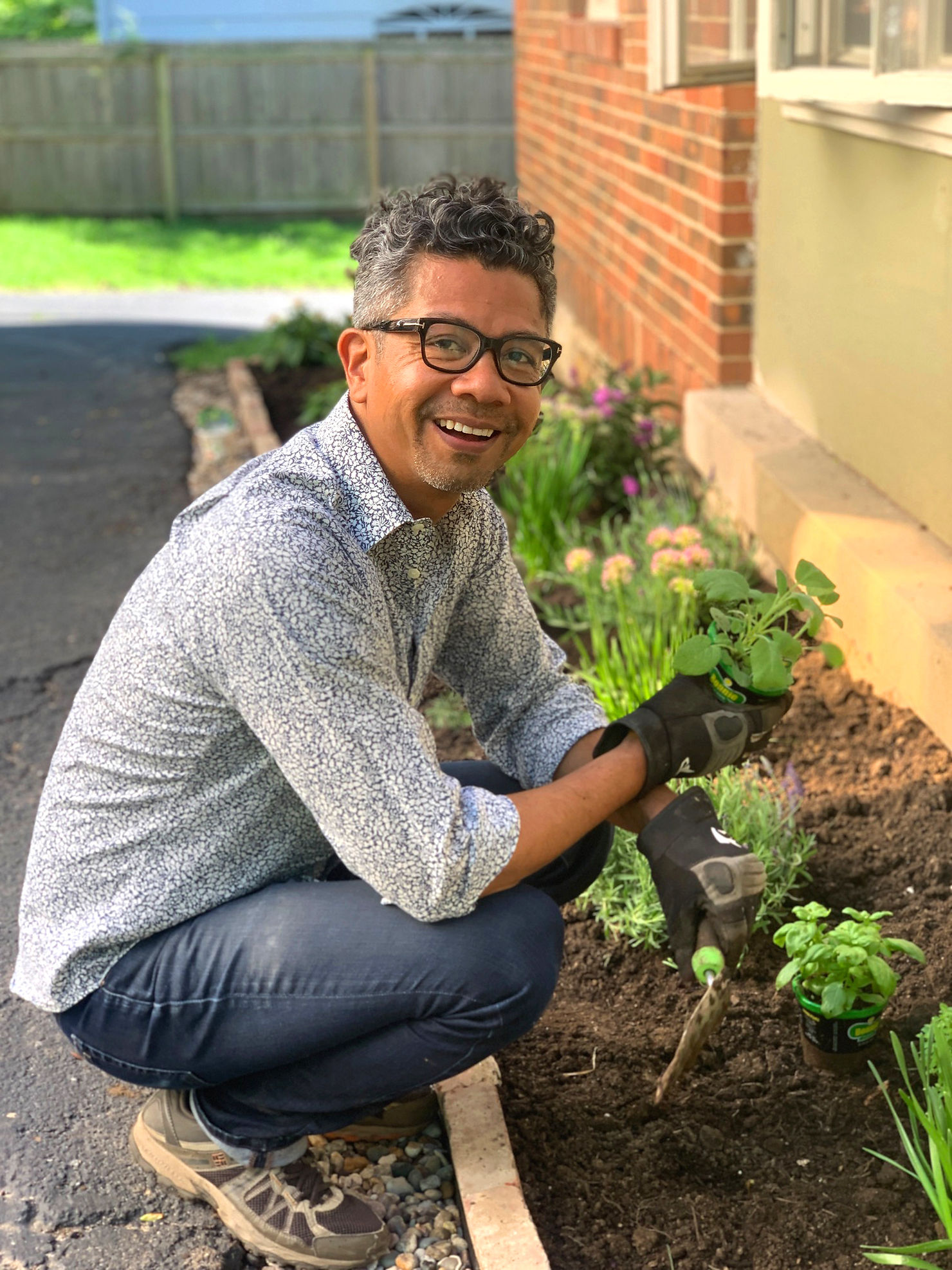 Man planting young flowers in a garden bed with fresh soil beside a brick home, wearing gardening gloves and holding a small plant outdoorsMan planting young flowers in a garden bed with fresh soil beside a brick home, wearing gardening gloves and holding a small plant outdoors