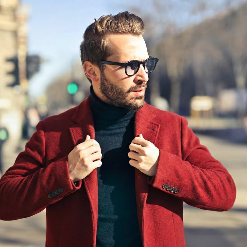 Man wearing sunglasses and a red coat adjusting his collar outdoors on a sunny day.