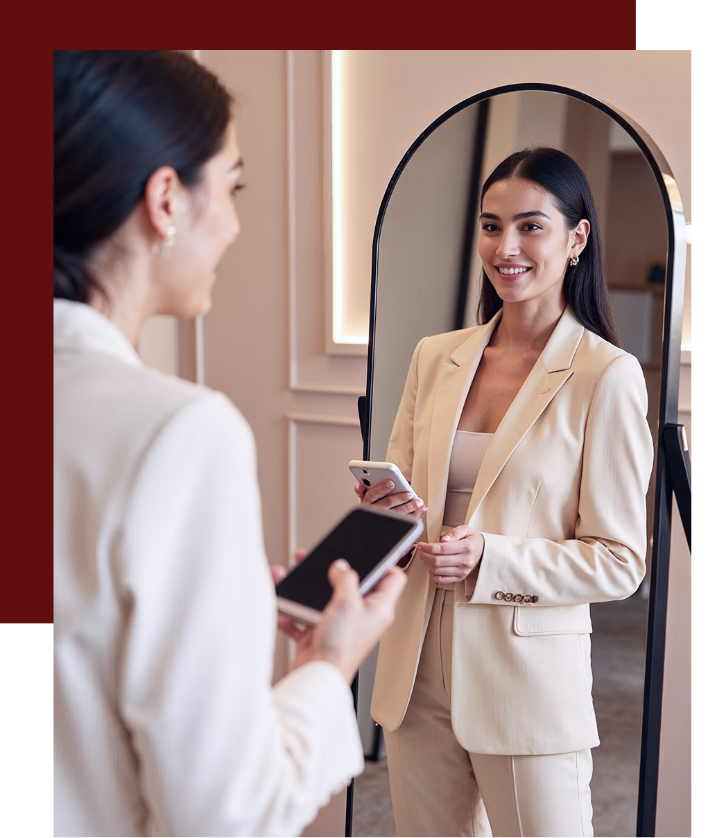 Smiling woman in beige suit holding a phone and looking at herself in a floor mirror.
