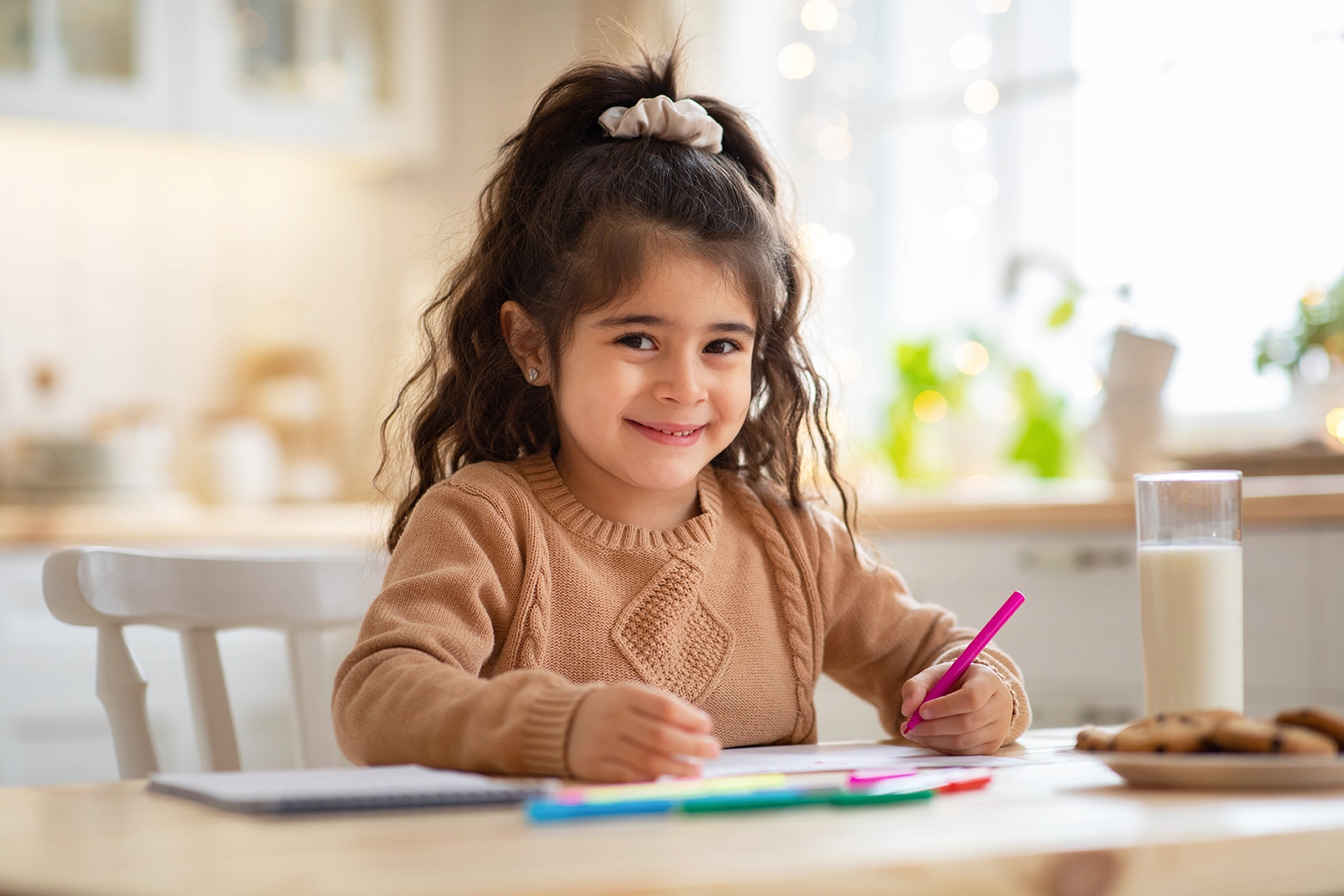 A young child smiling at the camera while drawing with colored pencils.