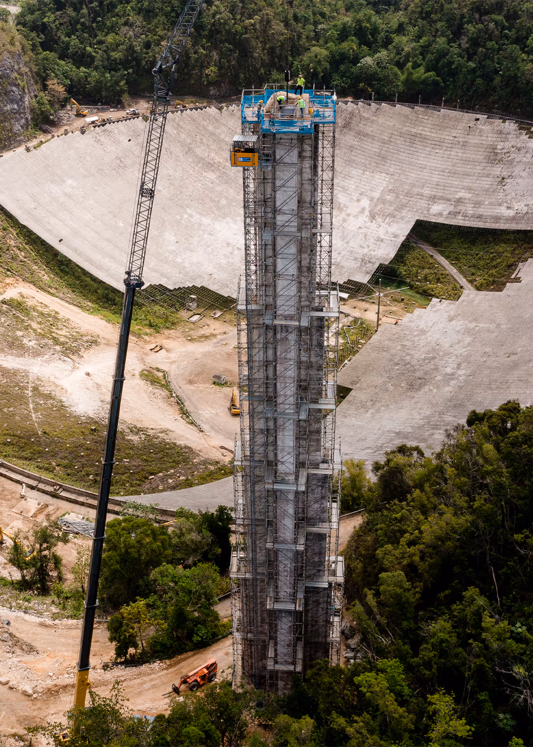 Aerial view of scaffolding tower restoration at Arecibo Observatory in Puerto Rico, with workers on top platform and crane lifting materials inside the iconic radio telescope dish.