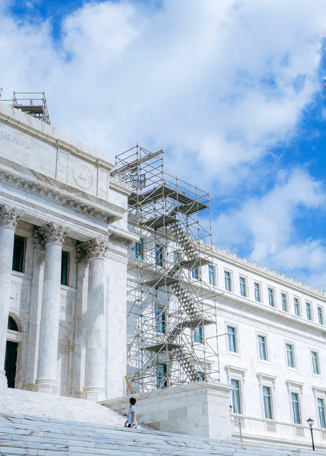 Side view of Puerto Rico Capitol building in San Juan with exterior scaffolding for restoration, marble columns and steps visible under blue sky.