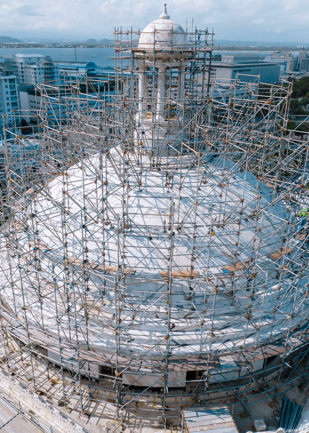Close-up aerial view of Puerto Rico Capitol dome in San Juan fully surrounded by scaffolding for structural restoration, overlooking city skyline and ocean.