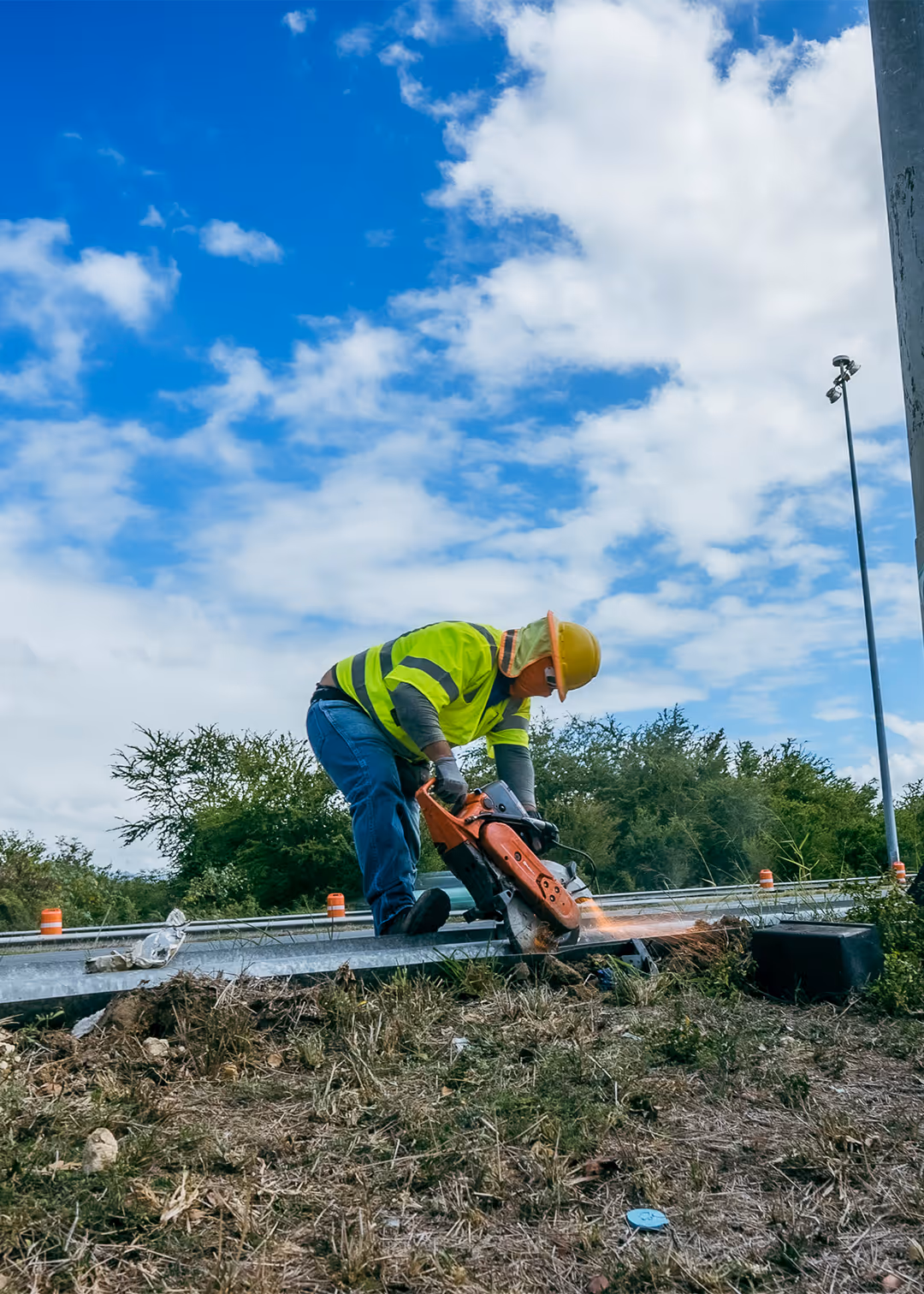 Construction worker in Puerto Rico cutting metal guardrail with power saw during road safety project, wearing high-visibility vest and protective gear under blue sky.