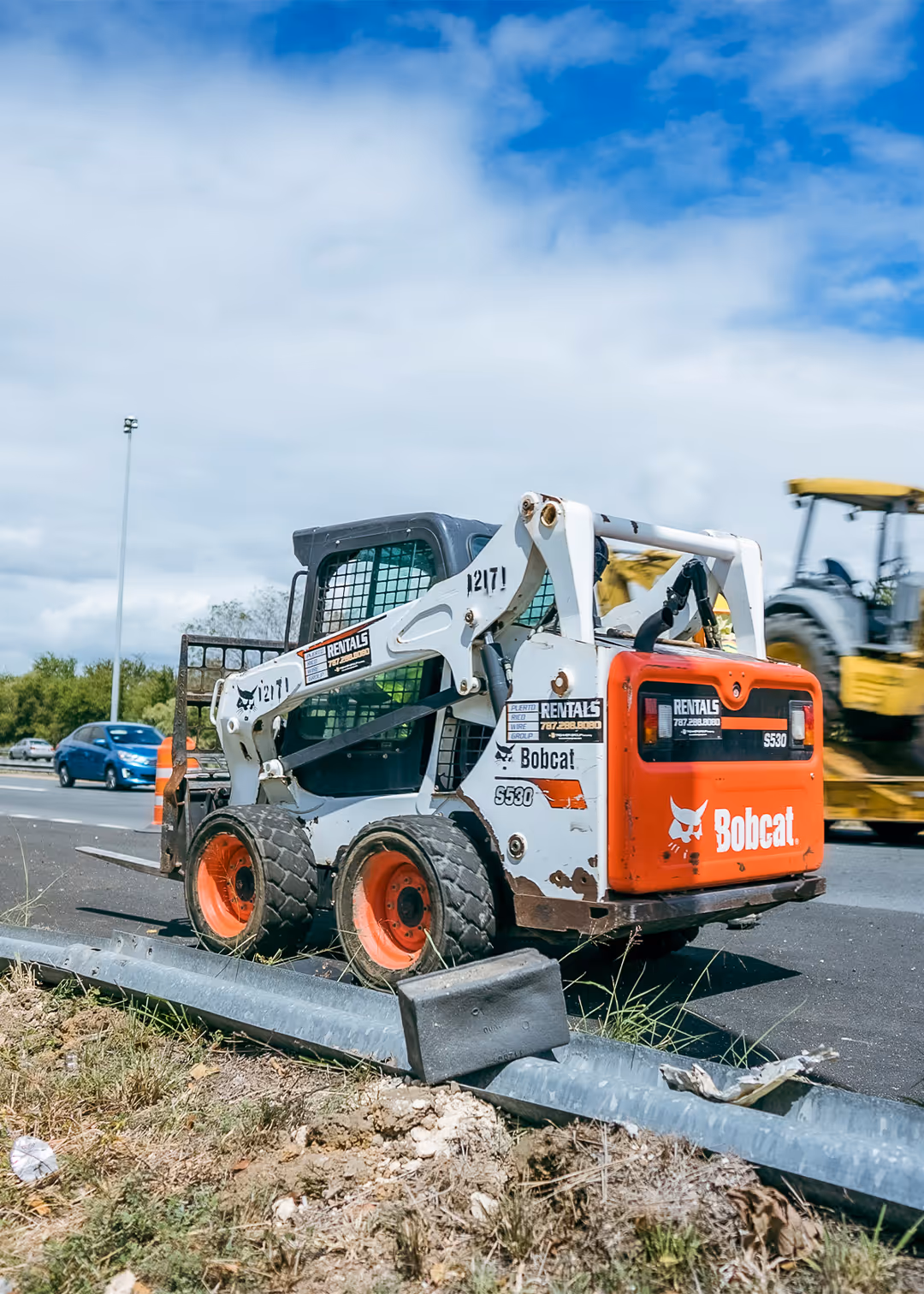 Bobcat skid steer loader with rental signage working on highway guardrail construction project in Puerto Rico, alongside heavy machinery and traffic.
