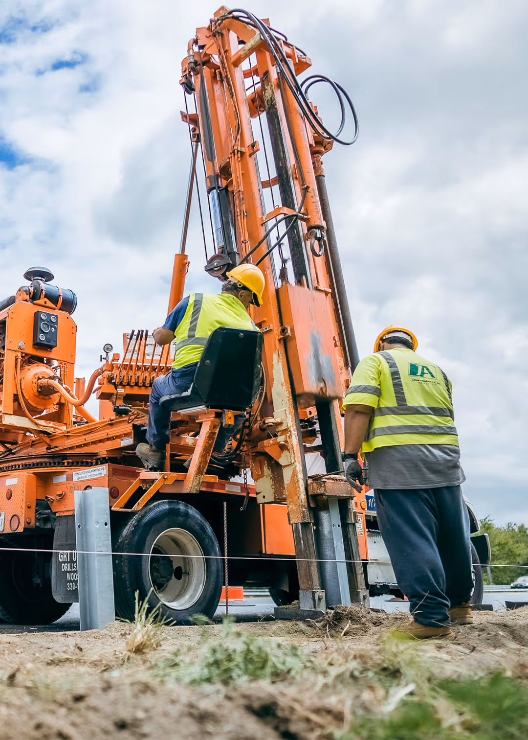 Construction workers operating heavy drilling machine to install guardrail posts during highway safety project in Puerto Rico, wearing safety vests and helmets.
