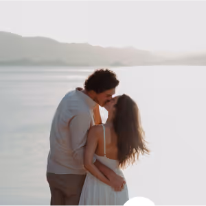 Couple embracing and kissing by a calm waterfront with mountains in the background during daylight.