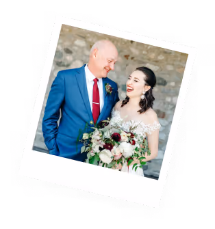 Bride in white dress holding bouquet standing next to man in blue suit and red tie against a stone wall background.