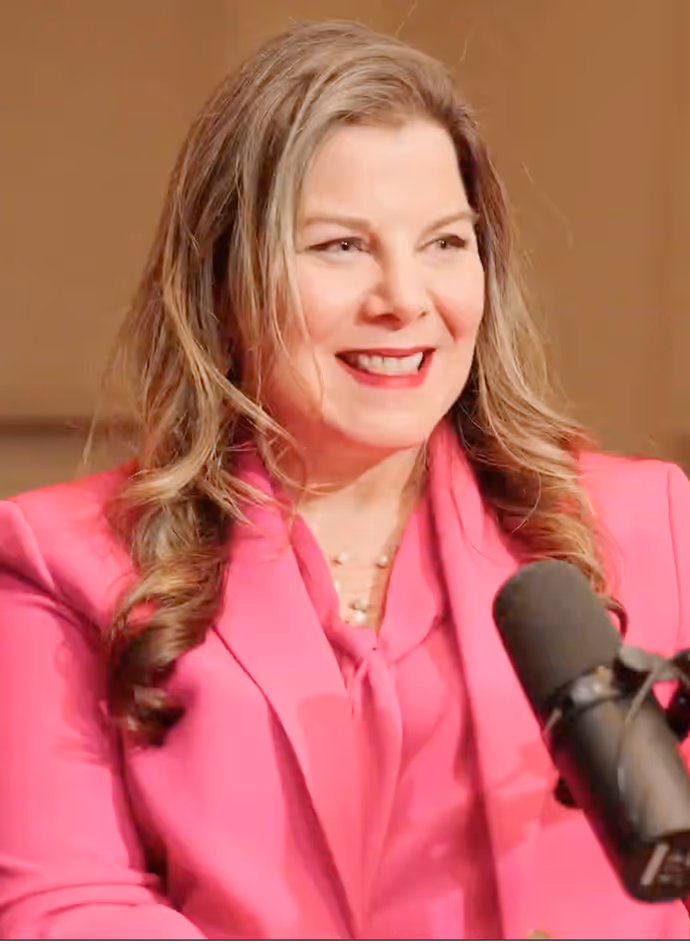 Woman with long wavy hair wearing a bright pink blazer, smiling while speaking into a microphone.