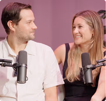 Man and woman smiling and looking at each other while sitting in front of microphones in a recording setting.