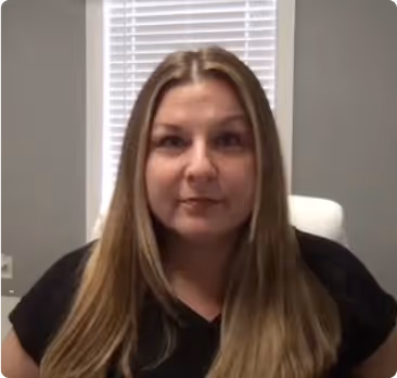 Woman with long straight hair wearing a black shirt, sitting in front of a window with blinds in a gray room.