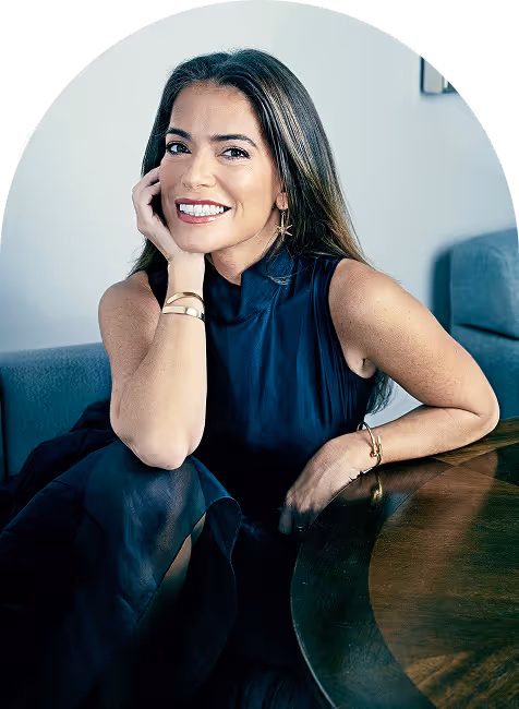Smiling woman with long hair wearing a sleeveless black dress, sitting at a wooden table with her hand resting on her cheek.