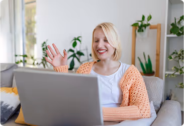 Smiling woman in orange sweater waving while video chatting on laptop in bright living room.
