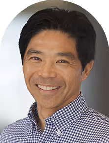 Smiling man with short black hair wearing a blue and white checkered shirt against a neutral background.