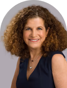 Smiling woman with curly brown hair wearing a sleeveless navy blue top and two layered necklaces.