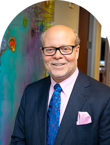Smiling bald man wearing glasses, a dark suit, blue patterned tie, pink shirt, and matching pink pocket square standing indoors.