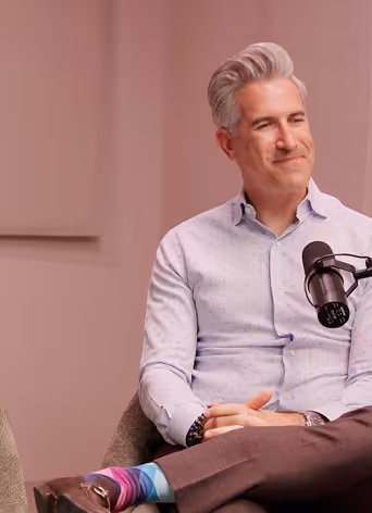 A man with gray hair in a light blue shirt sitting with crossed legs and a microphone on a stand in front of him.
