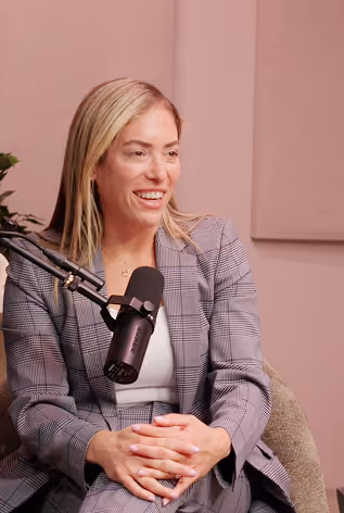 Smiling woman with blonde hair in a plaid suit sitting and speaking into a microphone in a podcast or interview setting.