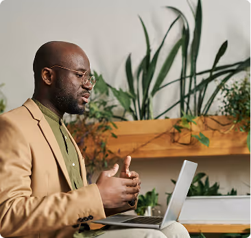 Man in beige jacket and glasses gesturing while talking on a laptop in a plant-decorated room.