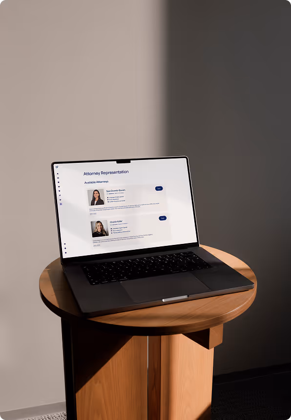 Open laptop displaying a webpage titled 'Attorney Representation' with profiles of two female attorneys on a round wooden table against a two-tone gray wall.