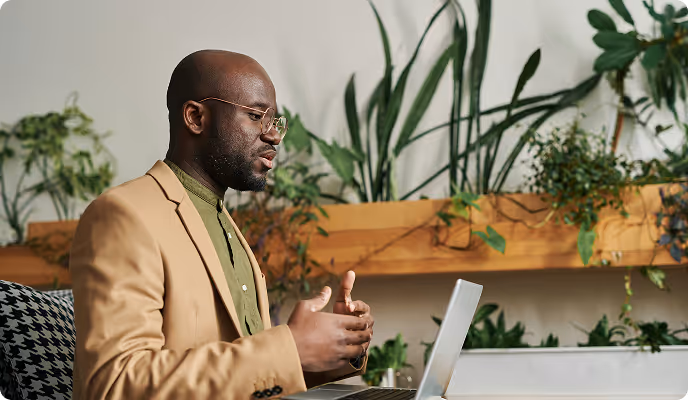 Man in glasses and a brown blazer speaking while looking at a laptop with green plants in the background.