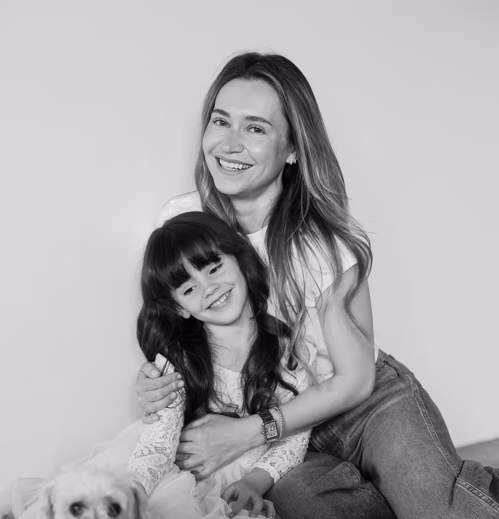 Smiling woman with long hair hugging a smiling young girl with bangs, seated together with a small dog in front of them.