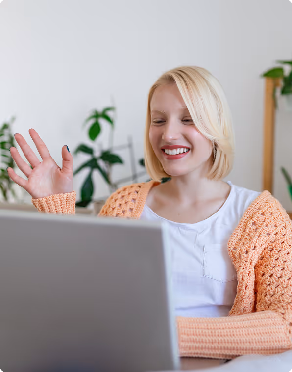 Smiling woman wearing a peach knitted cardigan waving while looking at a laptop screen.