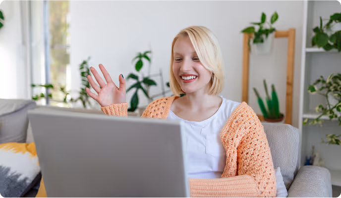 Smiling woman waving while video calling on a laptop in a cozy living room with plants.