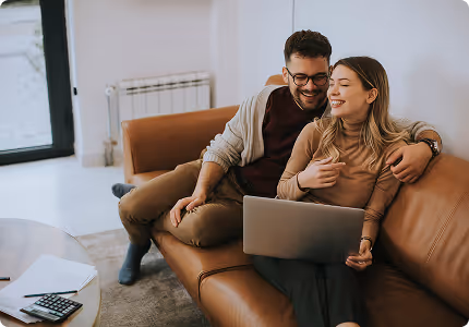 Smiling couple sitting on a brown leather couch, using a laptop together in a cozy living room.