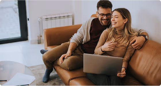 Smiling couple sitting on a brown leather couch using a laptop together in a bright living room.