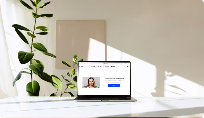 Laptop on a white desk displaying a video call interface with a woman smiling, next to a green potted plant in a sunlit room.