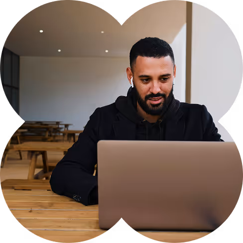 Man wearing wireless earbuds working on a laptop at a wooden table in a minimalist indoor space.