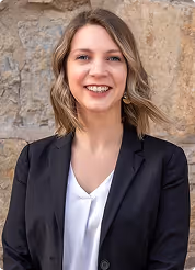 Smiling woman with shoulder-length blonde hair wearing a black blazer and white blouse, standing in front of a stone wall.