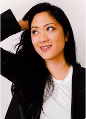 Smiling woman with long black hair wearing a black jacket and white top, looking to her left with one hand behind her head.