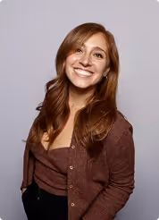 Smiling woman with long reddish-brown hair wearing a brown cardigan and top against a light gray background.