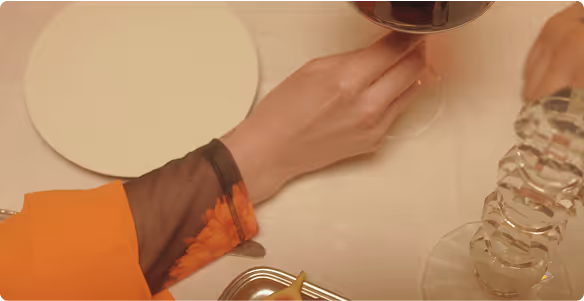 Hand holding a wine glass above a white tablecloth with an empty plate and stacked glass cups nearby.