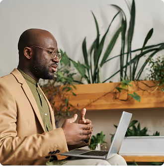 Man in glasses and beige blazer speaking while using a laptop indoors with plants in the background.