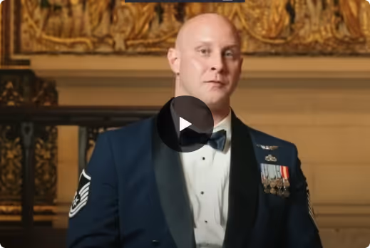 Man in formal military uniform with medals sitting indoors in front of ornate wall art.