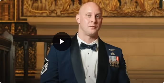 A bald man in a military dress uniform with medals and a bow tie standing indoors with ornate gold decorations in the background.