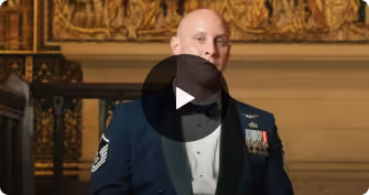 Man in military dress uniform speaking in an ornate room with a golden wall decoration.