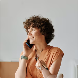 Smiling woman with curly hair wearing a peach blouse talking on a smartphone while holding eyeglasses.