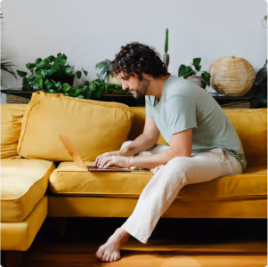 Man sitting barefoot on a yellow sofa, working on a laptop with green plants in the background.