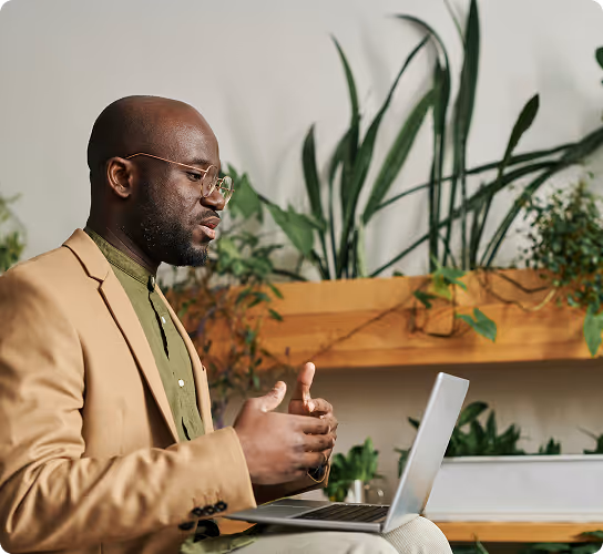 Man in tan blazer and glasses speaking while using a laptop with plants in the background.